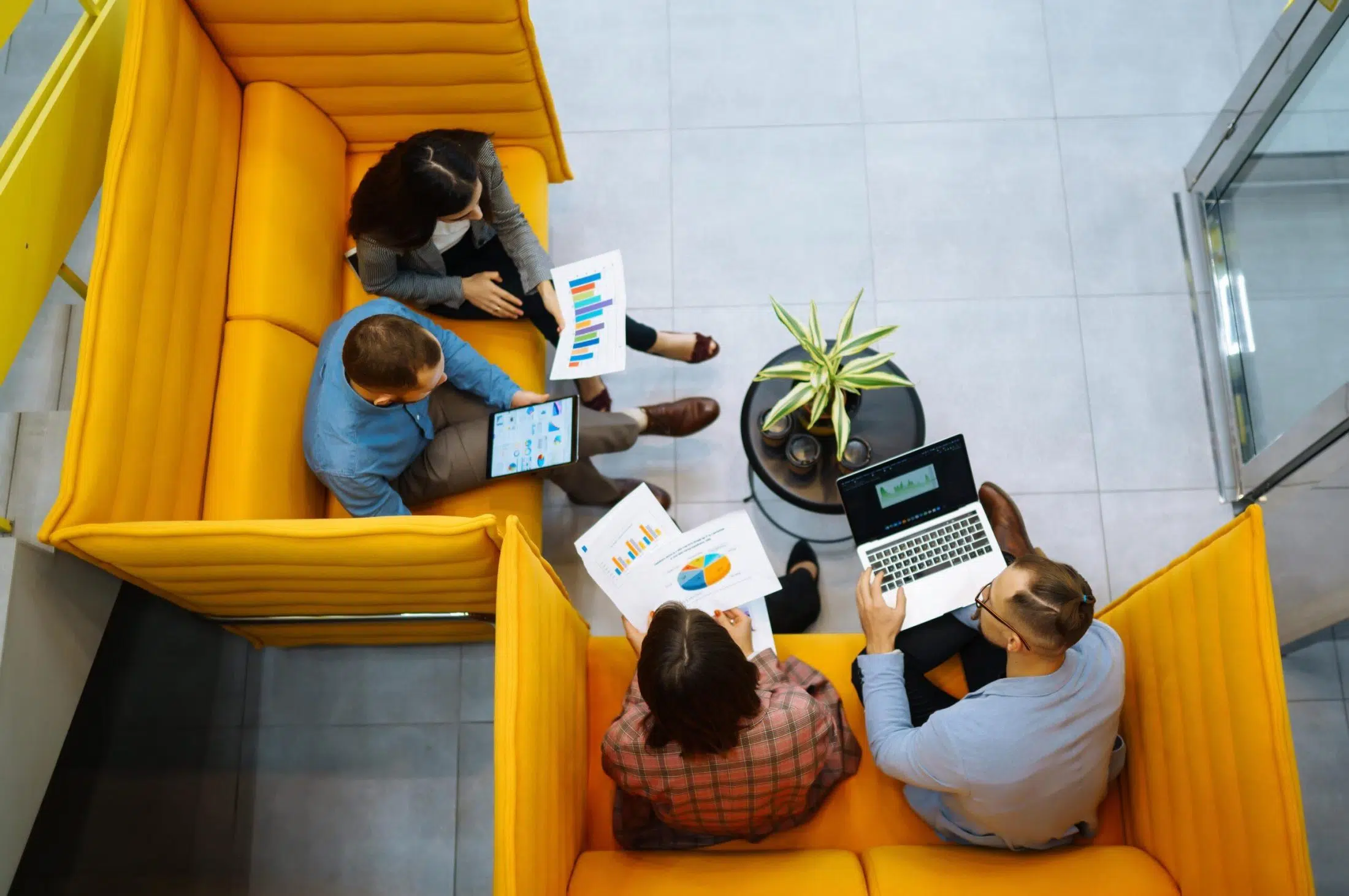 An overhead view of four people seated in a modern office lounge area with bright yellow, high-backed sofa seating arranged around a small round black table. One person holds a tablet displaying charts, while another holds printed sheets with colorful bar graphs and a pie chart. A third person works on a laptop showing a dashboard-style visualisation, and another person reviews printed data. A small green potted plant sits on the table. The floor is light grey tile, and the setting suggests a collaborative meeting focused on data analysis and reporting.