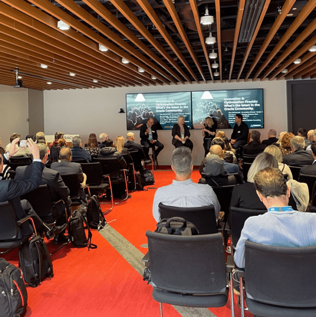Wide shot of a conference room during an Oracle event, showing a seated audience facing a four-person panel discussion at the front. The speakers sit on stools beneath large screens displaying “Innovation & Optimisation Fireside: What’s the latest in the Oracle Community,” with one speaker talking while others listen. The room features a red carpet, rows of chairs, and a wood-panelled ceiling.