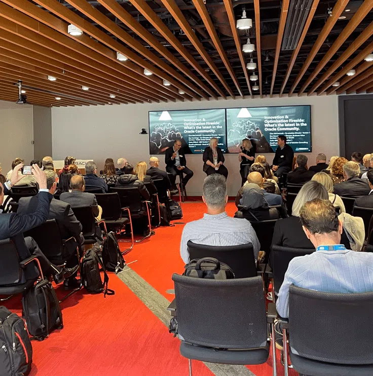 Wide shot of a conference room during an Oracle event, showing a seated audience facing a four-person panel discussion at the front. The speakers sit on stools beneath large screens displaying “Innovation & Optimisation Fireside: What’s the latest in the Oracle Community,” with one speaker talking while others listen. The room features a red carpet, rows of chairs, and a wood-panelled ceiling.