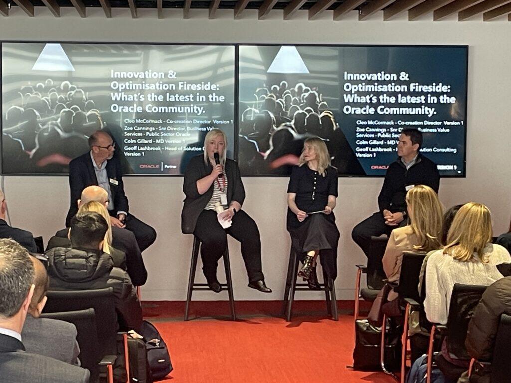 Panel discussion at an Oracle event, with four speakers seated on stools addressing an audience. A woman in the centre speaks into a microphone while others listen. Behind them, large screens display the session title: “Innovation & Optimisation Fireside: What’s the latest in the Oracle Community.”