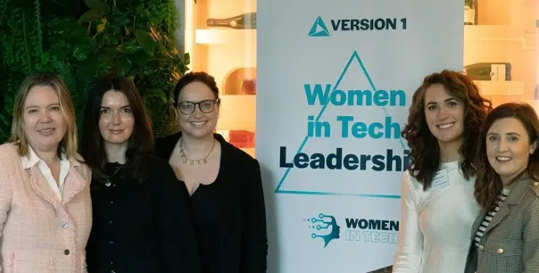 A group of five attendees standing side by side at a professional event, posing in front of a banner that reads “Version 1 Women in Tech Leadership.” The setting includes indoor greenery and warm lighting, with the banner and event space visible in the background.