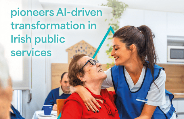 A promotional-style image set in a bright indoor care environment shows a healthcare worker in a blue uniform leaning over and embracing an elderly person seated at a table. The elderly person is wearing a red top and glasses, tilting their head upward toward the caregiver. In the background, another older individual is seated at a table with a cup, partially out of focus. The room appears clean and modern, with light-colored walls, a plant hanging from above, and a microwave visible on a shelf. Overlaid on the left side of the image in large teal text reads: &ldquo;pioneers AI-driven transformation in Irish public services.&rdquo; A stylised teal line graphic sits behind the caregiver and elderly person, adding a corporate branding element.