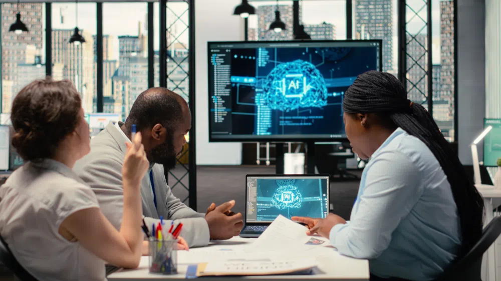 Three people sit around a table in a modern office with large windows and a city skyline visible in the background. In the center of the table, a laptop displays a visual interface featuring an abstract brain graphic labeled “AI,” surrounded by data elements. Behind the group, a large monitor shows a similar artificial intelligence-themed interface with code panels and a glowing brain visualization. Papers and pens are spread across the table, and the individuals appear to be discussing or reviewing the information on the screens, suggesting a collaborative meeting focused on AI or data analysis.Provide your feedback on BizChat