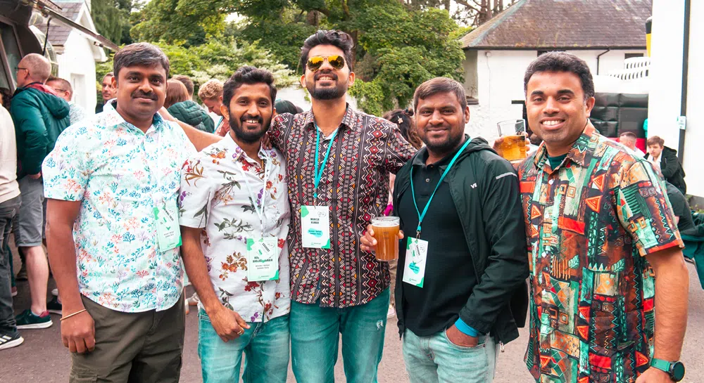 A group of five adults stand together outdoors at a casual networking or social event, posing for a photo. Each individual is wearing a lanyard with a visible event badge, suggesting a professional or careers-related gathering. Several of the individuals are dressed in patterned short-sleeve shirts, creating a relaxed and informal atmosphere. One individual near the center is holding a plastic cup filled with a drink. The group stands closely together in the foreground, facing the camera. Behind the group, more attendees are visible in a busy