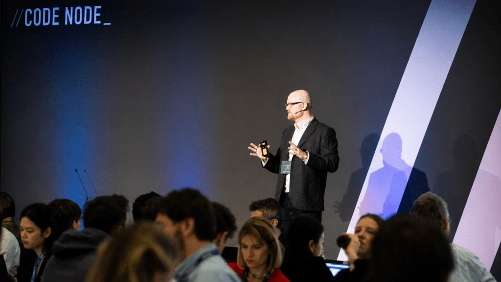 Conference speaker presenting on stage at a technology event, standing in front of a large dark backdrop with the text “//CODE NODE_”. Attendees are seated in the foreground, while the presenter speaks using a handheld device and headset microphone under blue and white stage lighting.