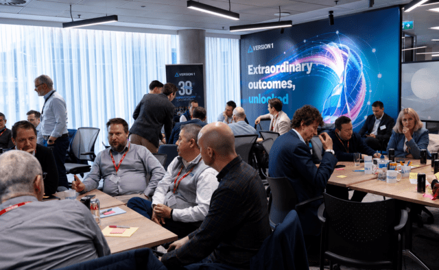 Business workshop with professionals seated in groups discussing ideas in a modern office, with a large “Extraordinary outcomes, unlocked” presentation backdrop.