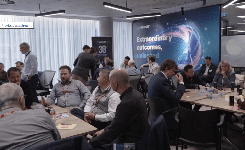 Business workshop with professionals seated in groups discussing ideas in a modern office, with a large “Extraordinary outcomes, unlocked” presentation backdrop.