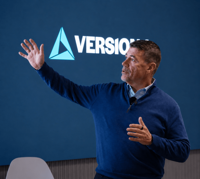 Man in a blue sweater speaking and gesturing with his hand in front of a screen displaying the Version 1 logo.