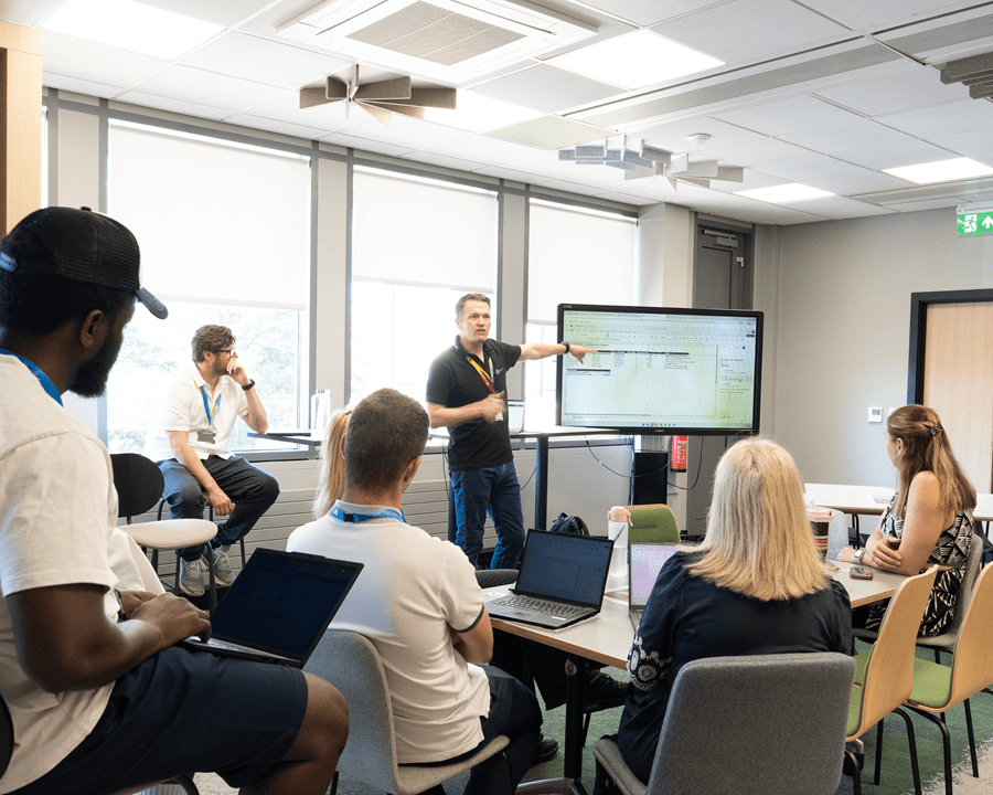 A group of people sit around tables in a modern meeting room while one presenter stands beside a large digital display, pointing to a spreadsheet during a workshop or team training session. Several attendees use laptops as they follow the presentation, with large windows providing natural light in the background.