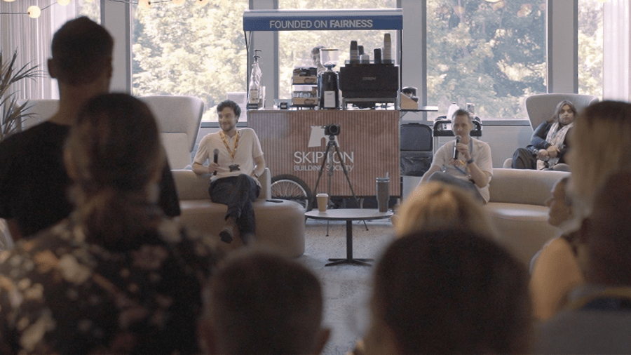 Audience members sit facing a panel discussion in a bright lounge-style event space. Two speakers are seated at the front near a small table and a branded coffee station displaying the text “SKIPTON Building Society” and “Founded on Fairness.” Large windows provide natural light, and a camera on a tripod is positioned between the speakers and the audience.