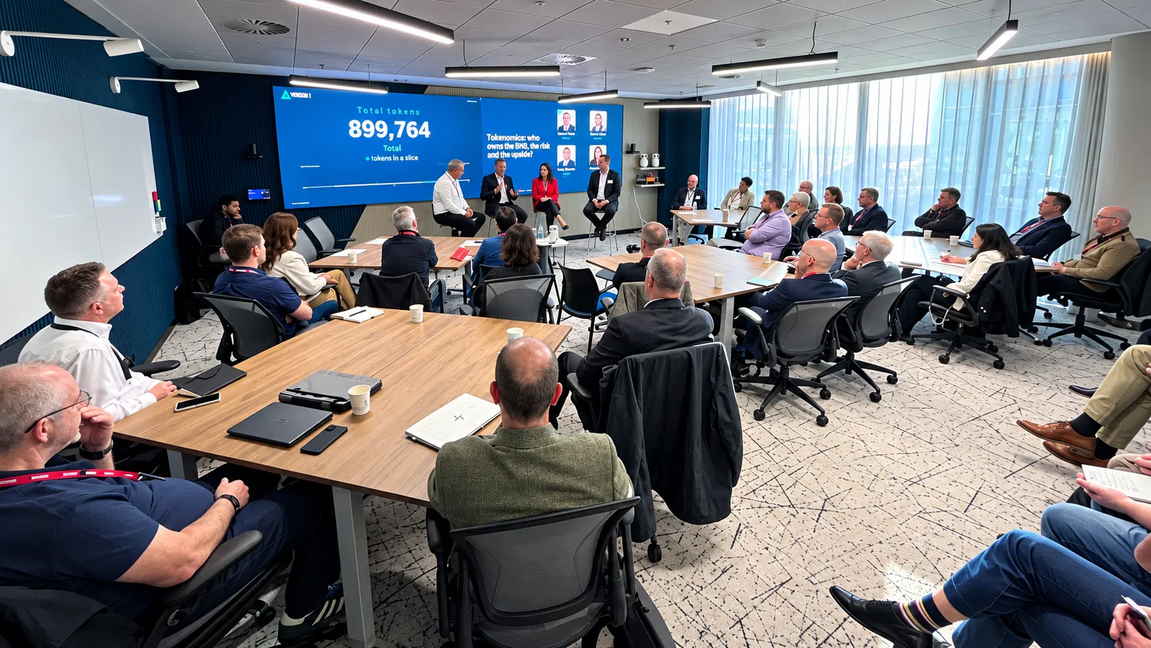 Professionals seated around tables in a conference room listening to a panel discussion, with a large screen displaying tokenomics data and presentation slides.
