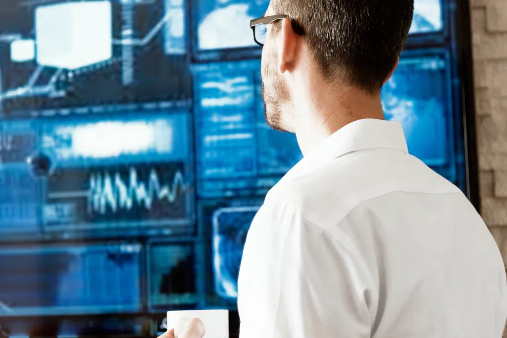 Person in a white shirt and glasses viewed from behind, looking at a large digital display wall showing blue-toned data visualizations, charts, and technology monitoring screens.