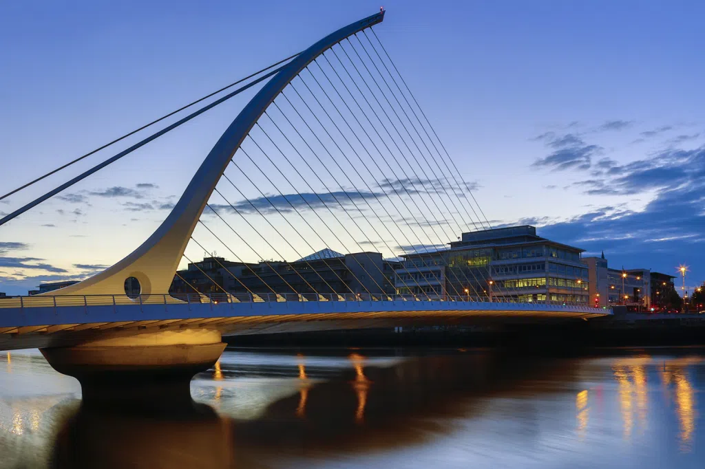 Samuel Beckett Bridge in Dublin, a modern cable-stayed bridge with a curved white harp-shaped pylon and cables spanning the River Liffey at dusk, with city lights reflecting on the water.