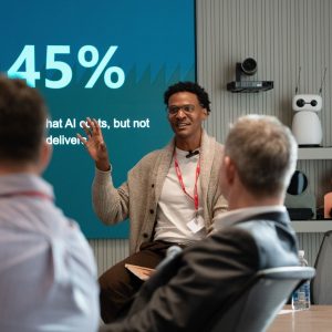 Speaker presenting at a Version 1 tokenomics event, seated in front of a large presentation screen displaying "45%". Attendees are visible in the foreground as the speaker addresses the audience in a modern conference room equipped with video conferencing technology.