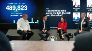 Panel discussion at a Version 1 tokenomics event featuring four speakers seated on stage in front of a large digital display. The screen shows token usage metrics, including "489,823 tokens used all time", alongside a presentation titled "Tokenomics: who owns the bill, the risk and the value?". Audience members are visible in the foreground as the panel discusses AI adoption, governance and value realisation.