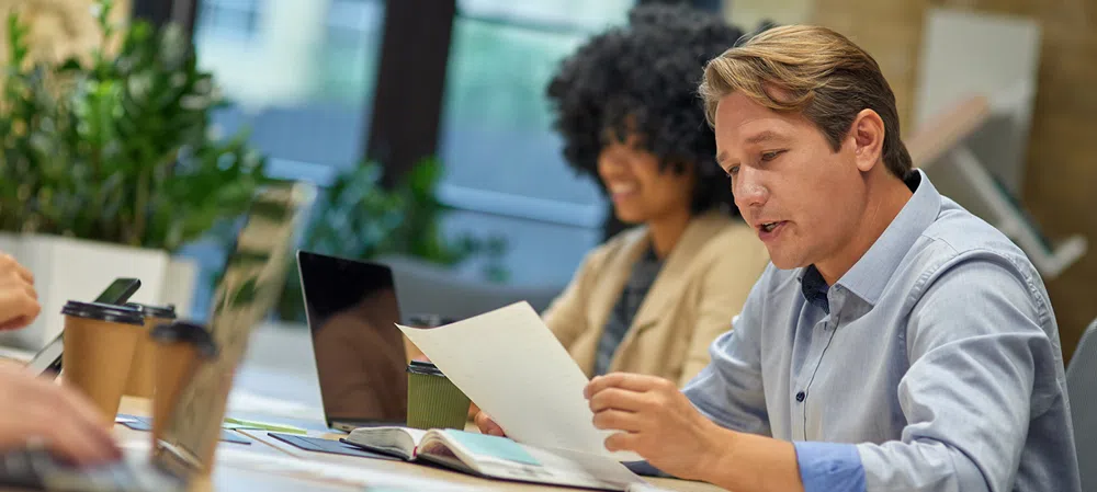Person reviewing a document at a meeting table with laptops, notebooks, and coffee cups in a modern office setting.