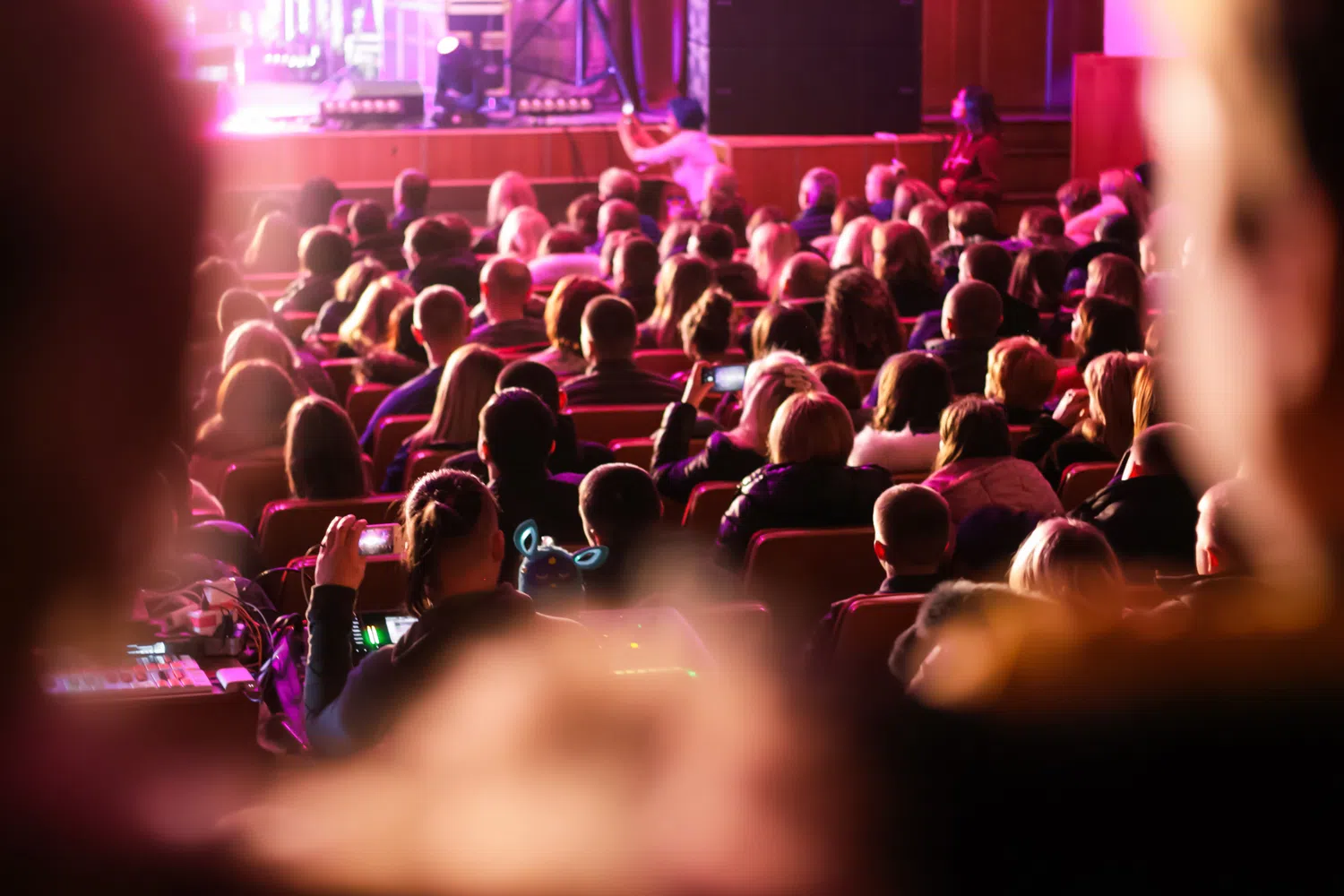 Audience seated in a theatre watching a live presentation or performance on a brightly lit stage, with purple and pink lighting illuminating the venue.