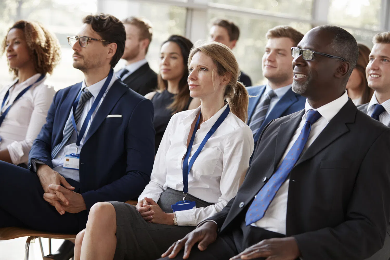 Business professionals seated in a conference or corporate seminar, listening to a presentation in a bright modern meeting space. Attendees wear business attire and event lanyards while facing a speaker outside the frame.