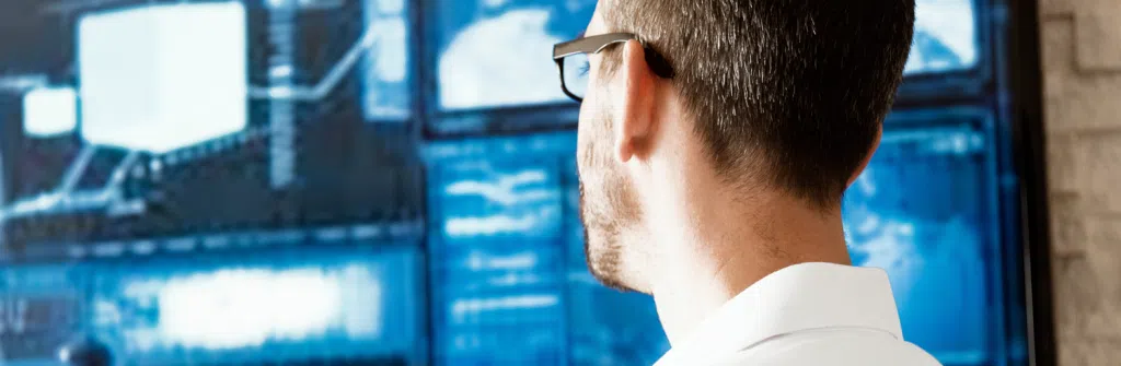 Person in a white shirt and glasses viewed from behind, looking at a large digital display wall showing blue-toned data visualizations, charts, and technology monitoring screens.