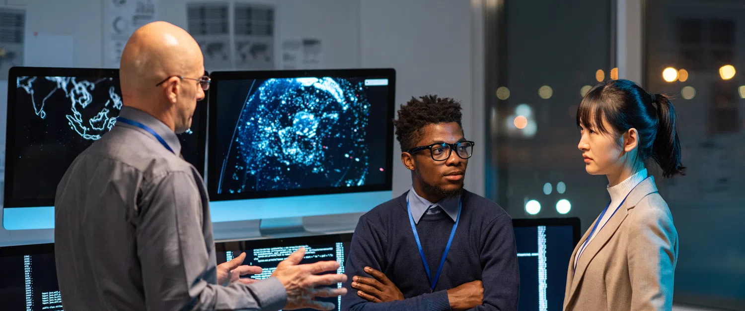 Three technology professionals wearing business attire and ID lanyards discuss AI and data analytics in a modern office, with multiple monitors displaying digital maps, data visualisations and computer code in the background.