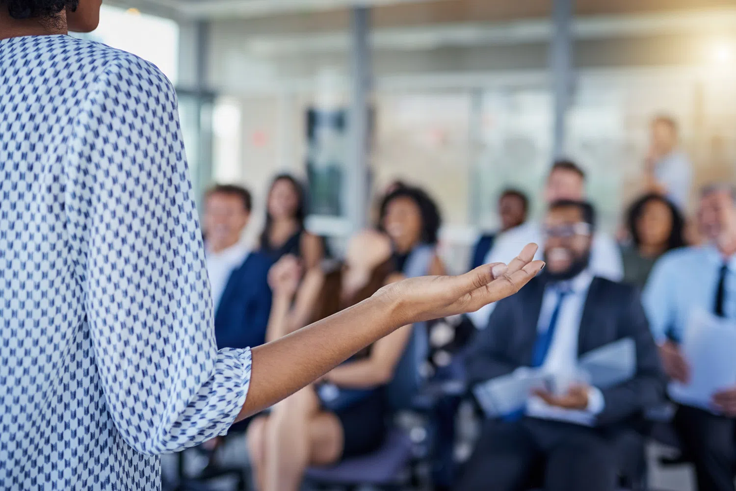 Person speaking to a group in a modern office or conference setting, viewed from behind with an outstretched hand gesturing toward an audience seated in the background. The audience is softly blurred, emphasizing the presenter and the act of public speaking, training, or business communication.