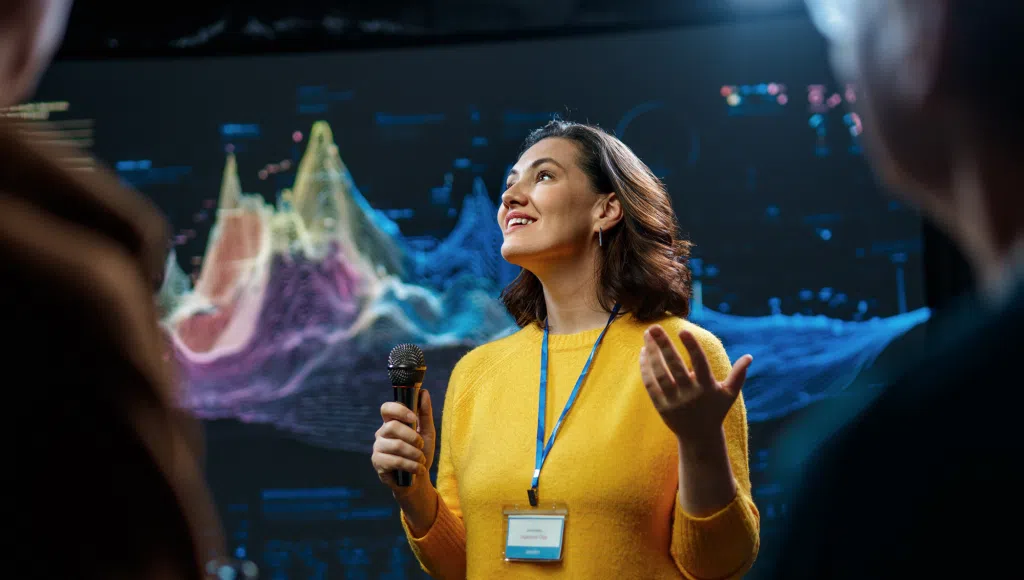 Presenter speaking into a microphone at a technology conference, standing in front of a large digital data visualisation display. The speaker wears an event badge and addresses an audience while colourful analytics graphics appear on a screen in the background.