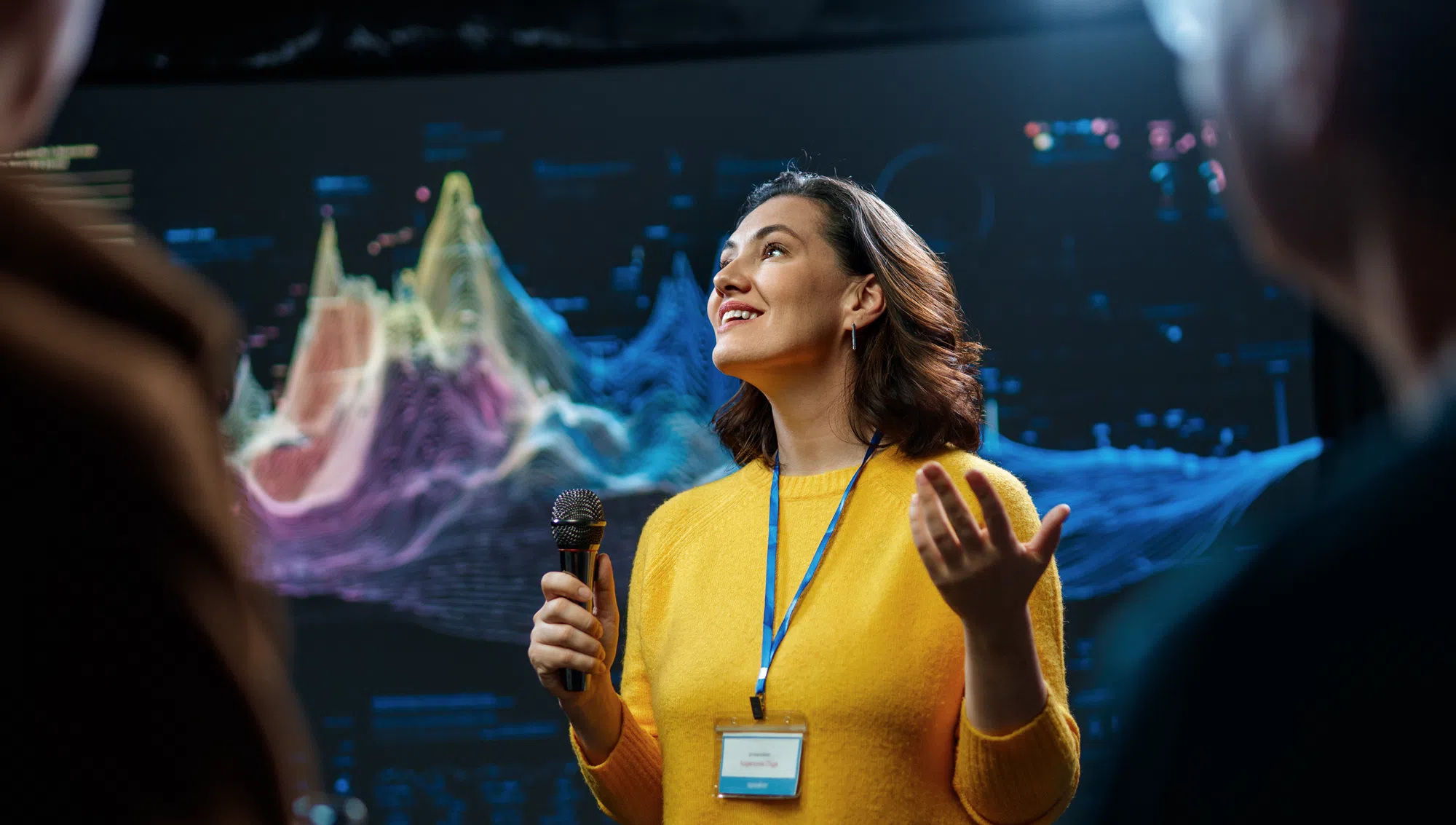 Presenter speaking into a microphone at a technology conference, standing in front of a large digital data visualisation display. The speaker wears an event badge and addresses an audience while colourful analytics graphics appear on a screen in the background.