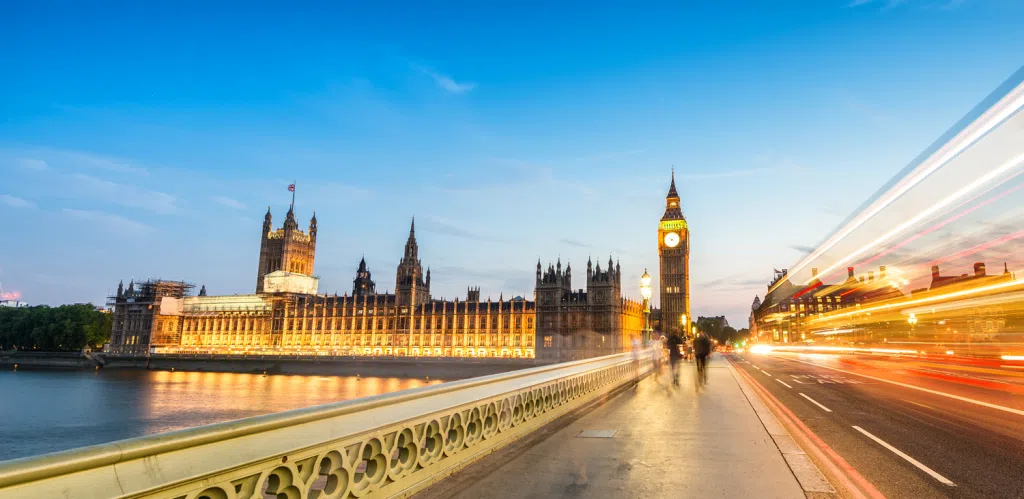 Illuminated Houses of Parliament and Big Ben in London at dusk, viewed from Westminster Bridge, with the River Thames in the foreground and blurred pedestrians and traffic creating light trails along the bridge.