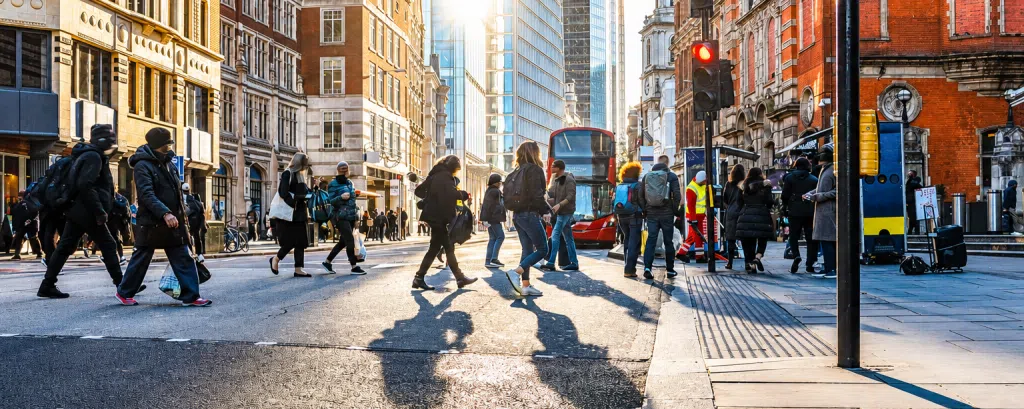 People crossing a busy London city centre street with a red double-decker bus and commercial buildings, representing urban infrastructure, public services and business transformation in the UK.