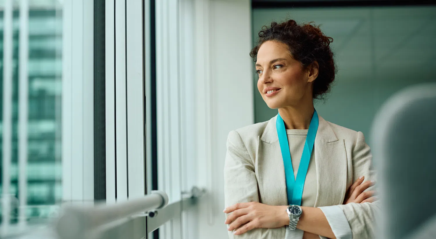 Professional standing beside a large office window with arms crossed, wearing a light blazer and teal lanyard, looking out across a modern city office environment with natural daylight.