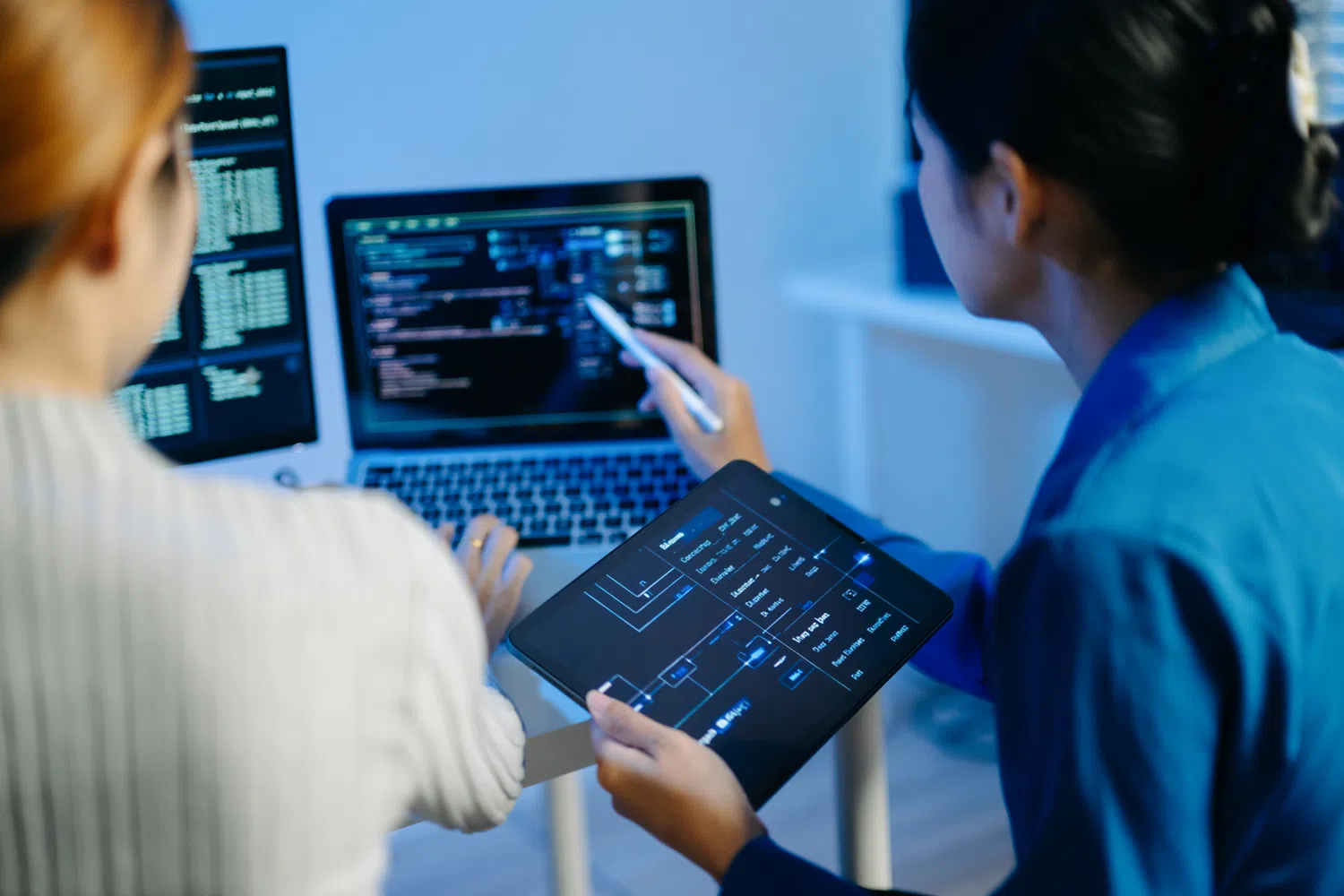 Two professionals collaborating on software development, reviewing code and system architecture on multiple screens and a tablet in a modern technology workspace.