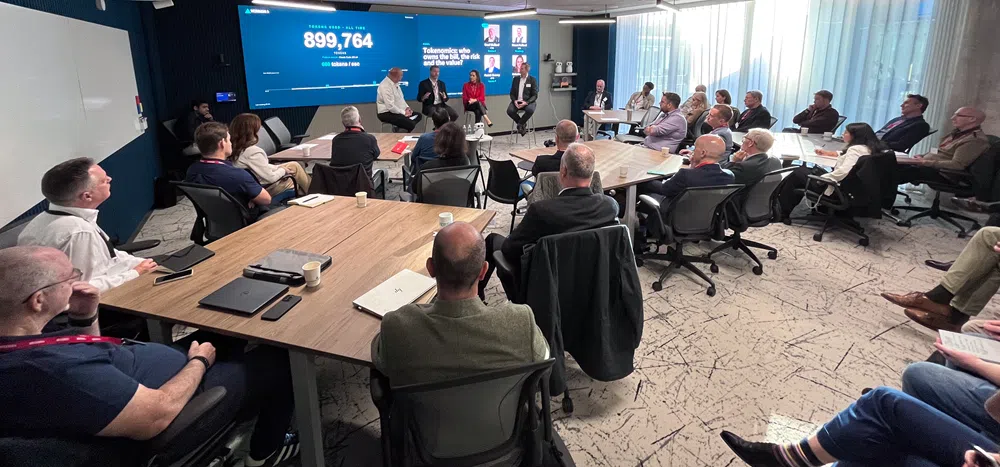 Professionals seated around tables in a conference room listening to a panel discussion, with a large screen displaying tokenomics data and presentation slides.