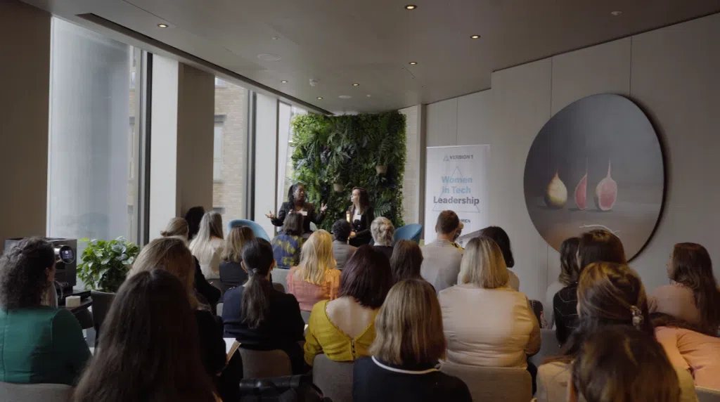 Audience seated in a modern conference room listening to a panel discussion, with two speakers at the front, a living green wall backdrop, large floor-to-ceiling windows, and a Version 1 Women in Tech Leadership banner displayed beside the stage area. A circular artwork hangs on the wall to the right, while natural light fills the room.